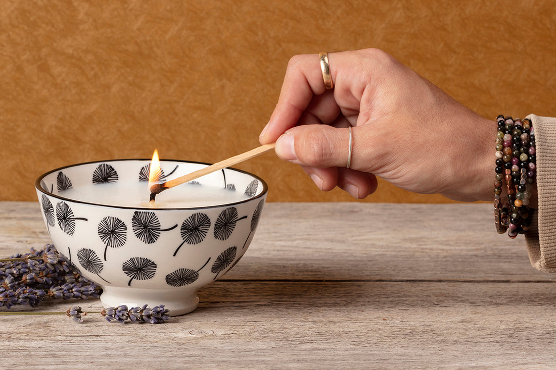 Hand lighting a candle with a wooden wick, surrounded by lavender flowers on a wooden surface.
