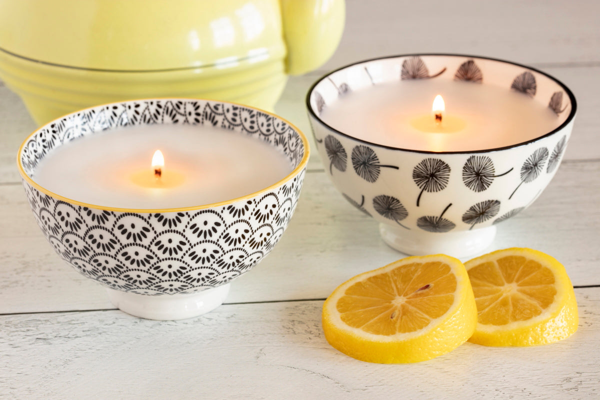 Two ceramic bowl candles, one with a lit white candle, the other is empty, with black and white floral patterns. Lemon slices are also present on the surface.