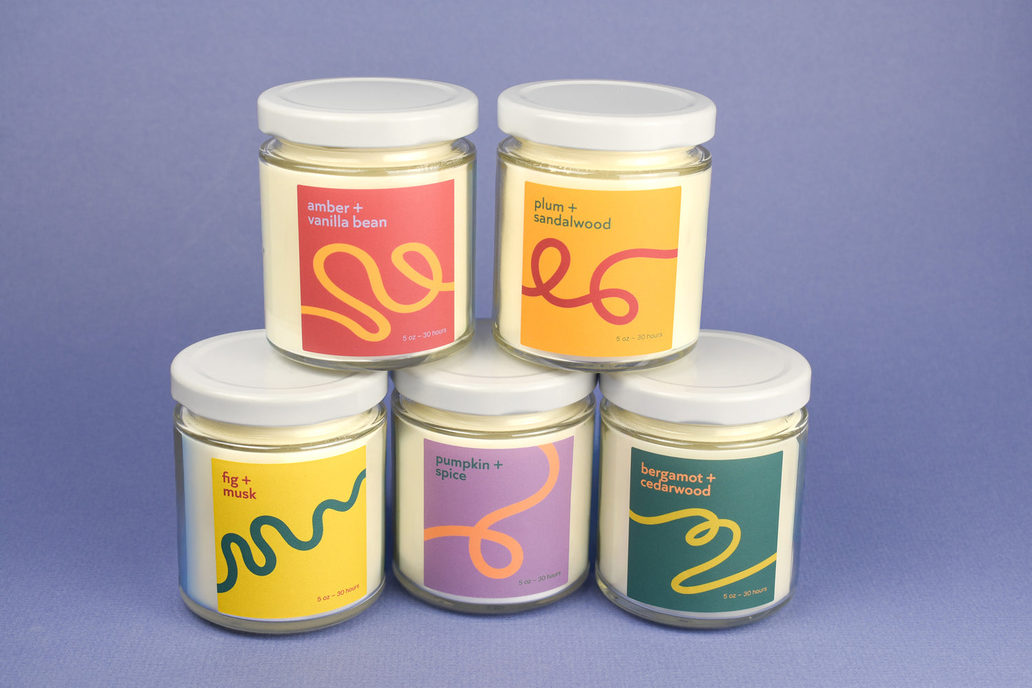 Five glass jar candles with white lids are arranged in a pyramid on a lavender background. Each candle has a different colorful label with abstract wavy line art and scent names including amber + vanilla bean, sandalwood, fig + musk, pumpkin + spice, and bergamot + butterwood.