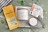 A styled flat-lay of three artisanal food and beverage products on a light sage green surface with scattered glass pebbles. On the left is a yellow Moonstruck milk chocolate salted toffee box, in the center a white candle jar labeled &