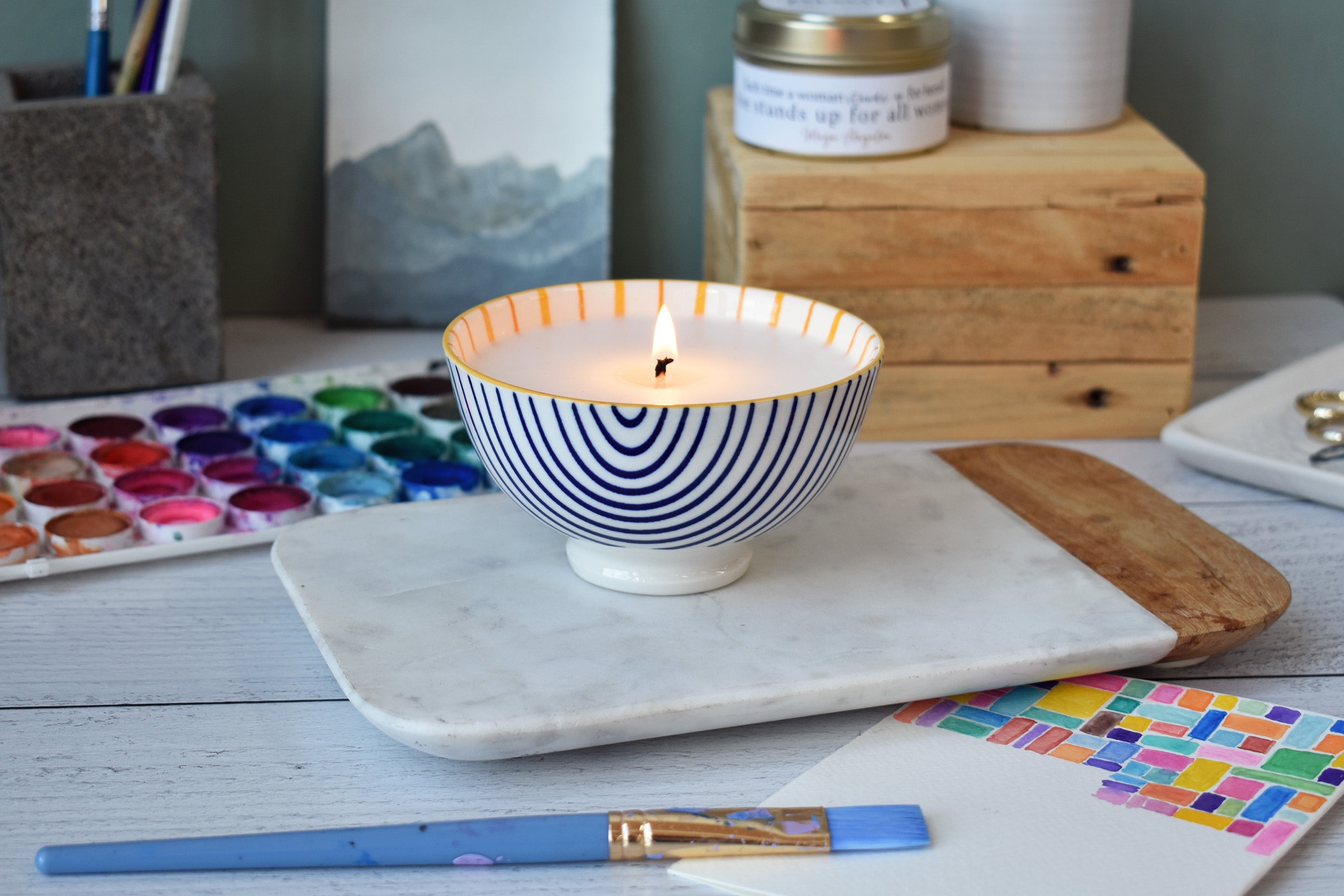 Candle in a decorative bowl on a marble tray with art supplies in the background